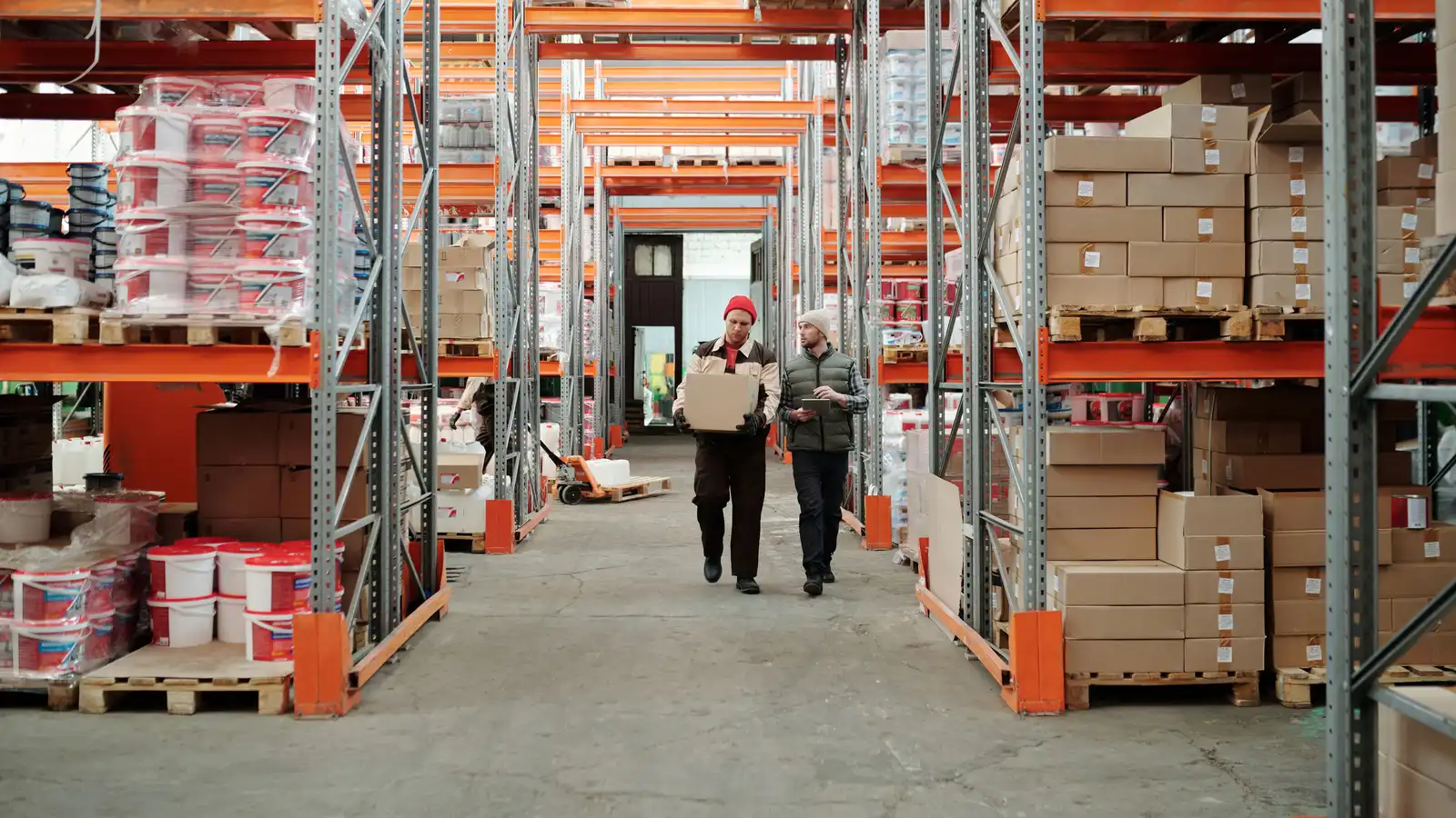 Two warehouse workers walking up an aisle of racked stock — one carrying a carton, the other checking a tablet.