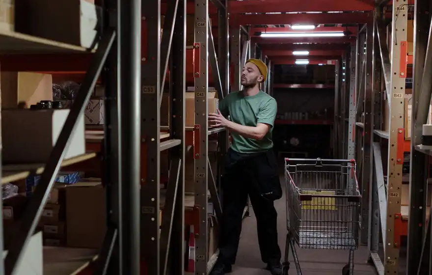 A store-room worker picking supplies from shelving beside a stock trolley.