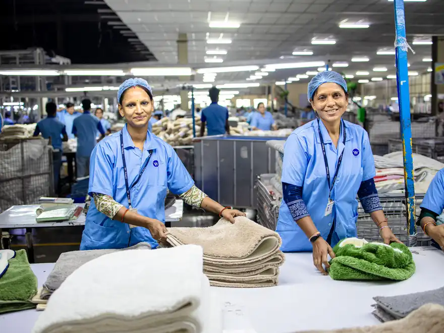 Two workers in uniform folding and stacking finished textiles at a bench, with the rest of the factory floor at work behind them.