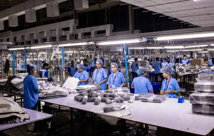 Workers in uniform folding and packing goods along benches on a factory production floor.